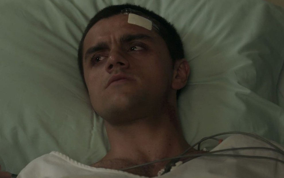O ator Felipe Simas está deitado em uma cama de hospital com expressão de dúvida em cena de Salve-se Quem Puder