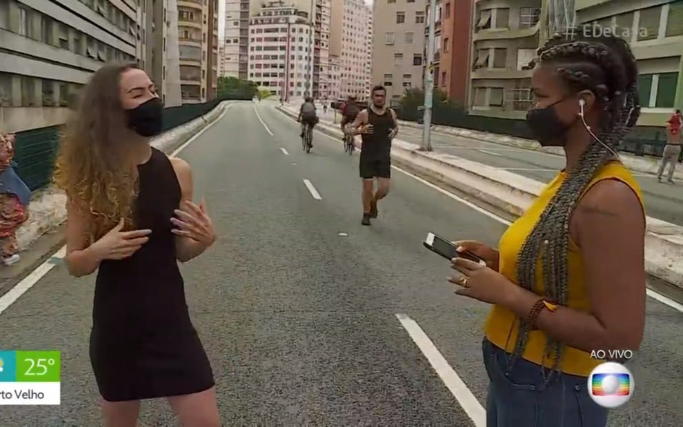 Mulher loira, de vestido preto, conversa com outra mulher negra, de blusa amarela e calça jeans em uma rua a céu aberto