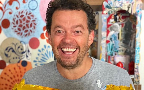Fernando Rocha sorri para foto e usa camiseta cinza; ele está na frente de um fundo colorido