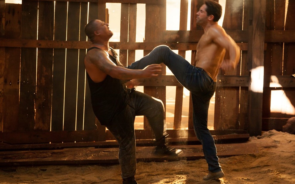 Sem camisa, ator Scott Adkins (à direita) chuta com a perna esquerda o peito do adversário