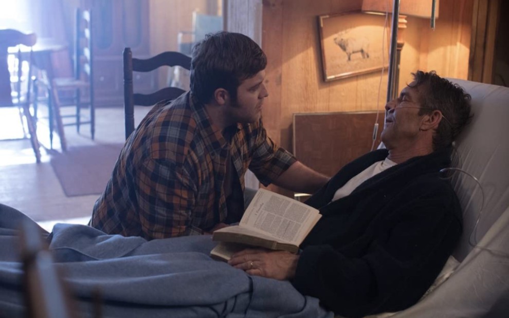 J. Michael Finley sentado com a mão apoiada em Dennis Quaid, que está deitado numa cama, aparentemente doente, com um livro em mãos
