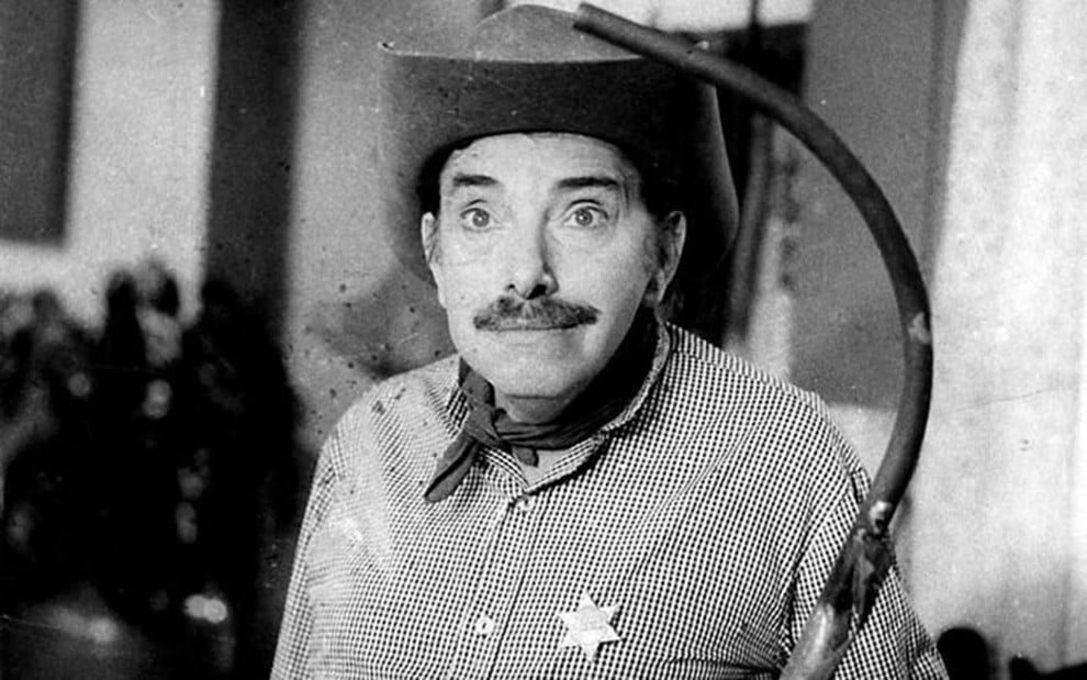 Mazzaropi em um de seus filmes, trajando chapéu, camisa, estrela de xerife e uma espingarda