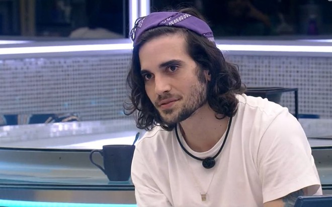 Fiuk olha para a esquerda usando uma camisa branca e uma faixa roxa em seu cabelo; ele está na cozinha do BBB21