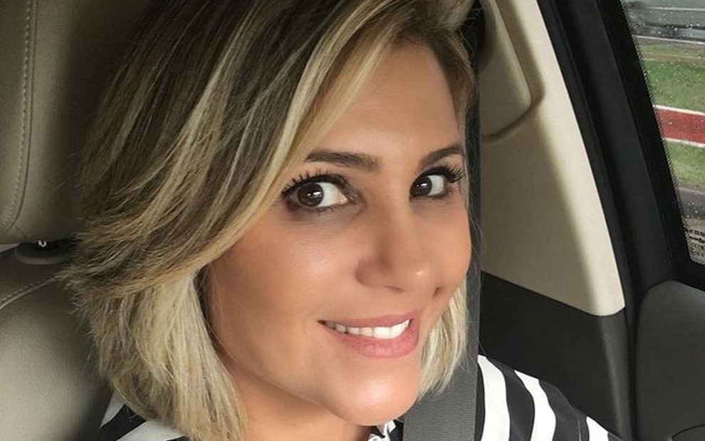 Flávia Freire sorridente, de cabelo curto com luzes, dentro do carro com blusa listrada de preto e branco
