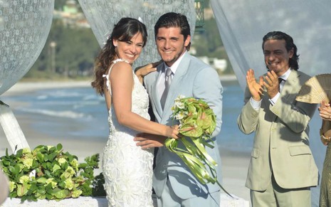 Os atores Daniela Escobar e Bruno Gissoni abraçados, sorriem, vestidos de noiva e noivo, ela com buquê em mãos, numa praia em cena de Flor do Caribe
