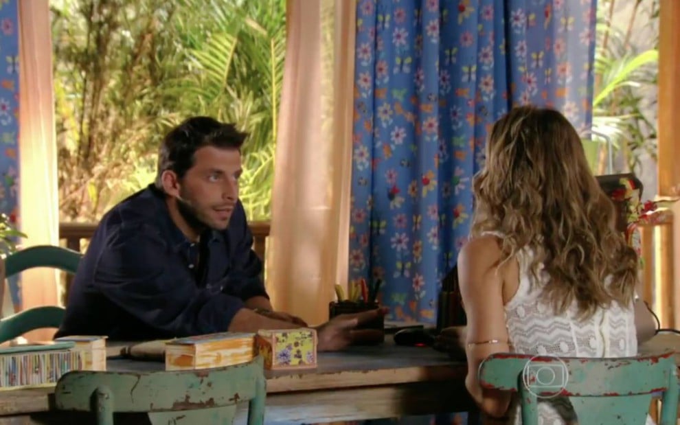 Os atores Henri Castelli e Grazi Massafera em cena de conversa, um em cada lado de uma mesa, em Flor do Caribe