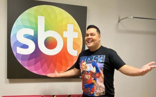 Everton Di Souza, o Fofoquito, aparece sorrindo ao lado de quadro com logotipo do SBT