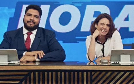 Os humoristas Paulo Vieira e Renata Gaspar numa bancada, cenário de telejornal do humorístico Fora de Hora