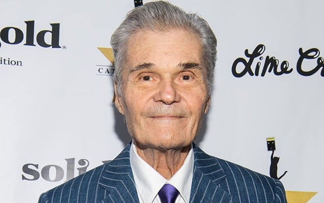 O ator Fred Willard sorri timidamente em frente a um painel de premiação