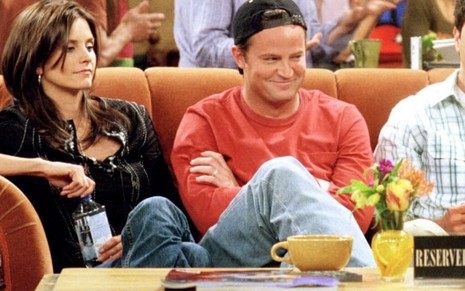 Sentados em um sofá, Courteney Cox olha para o lado esquerdo, Matthew Perry ri e David Schwimmer segura xícara em Friends