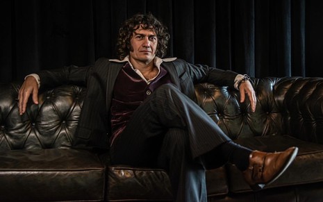 Gabriel Braga Nunes sentado em um sofá, com pernas cruzadas; ator posa para ensaio fotográfico de projeto musical
