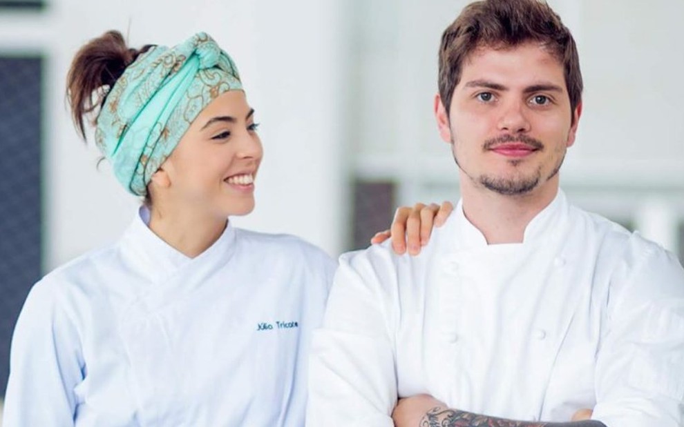 Os chefs de cozinha e noivos Júlia Tricate e Gabriel Coelho com uniformes brancos de cozinheiros, um ao lado do outro