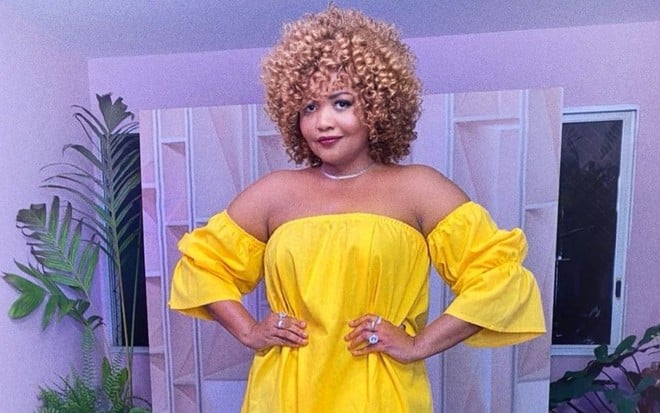 Gaby Amarantos com vestido amarelo, com mãos na cintura, em um cenário roxo