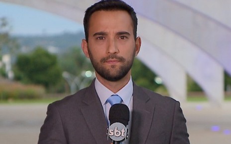 Galton Sé vestindo terno e gravata, e segurando microfone durante uma reportagem