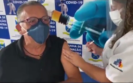 Galvão Bueno com manga levantada, máscara e óculos, recebe a vacina de profissional da saúde