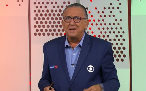 Galvão Bueno no estúdio da Globo com o terno azul marinho do uniforme; ele segura uma ficha com o logo da Globo e uma caneta