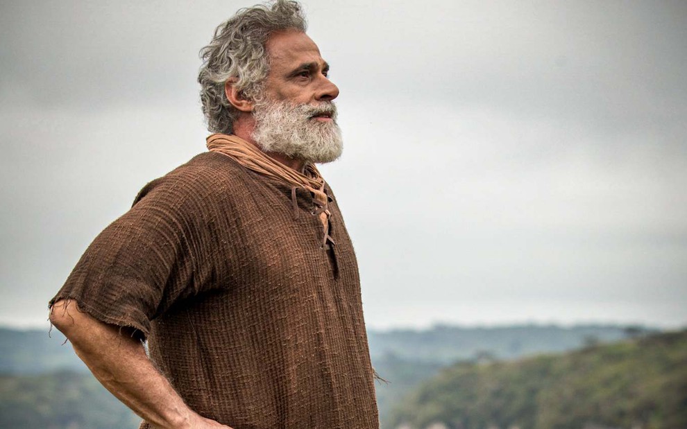 O ator Oscar Magrini em paisagem de campo, caracterizado como Noé, olhando para o horizonte em Gênesis