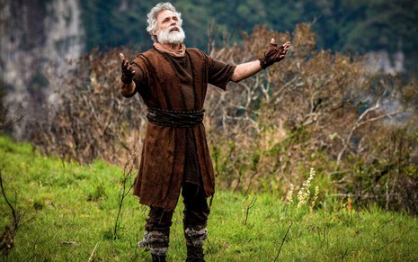 O ator Oscar Magrini em paisagem de campo, caracterizado como Noé, de braços abertos em cena de Gênesis