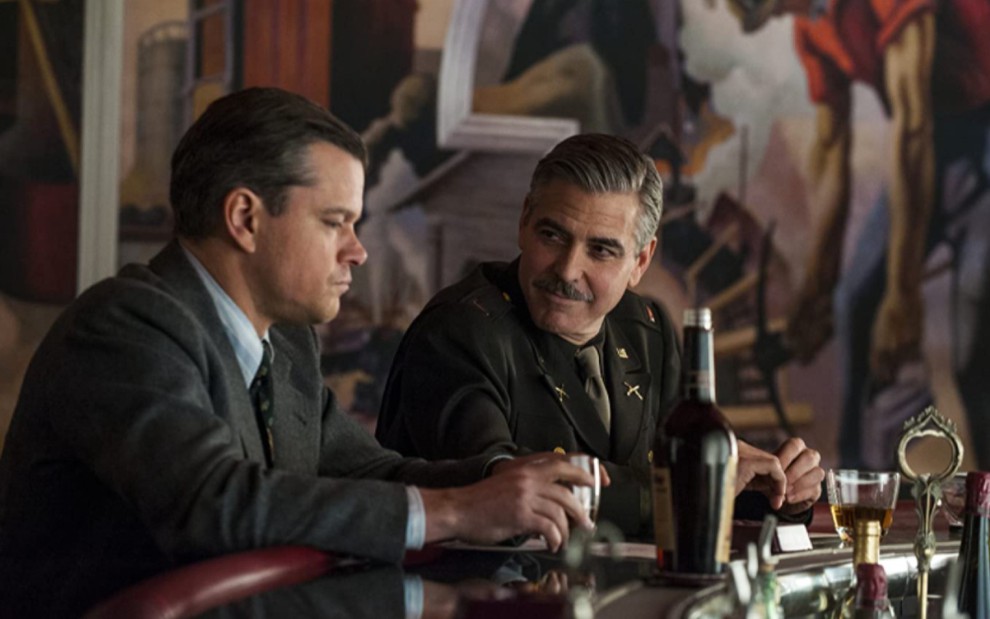 Matt Damon de perfil, mexendo em um copo, enquanto George Clooney olha para ele em Caçadores de Obras-Primas