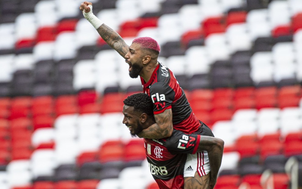 Gerson está de pé e carrega nas costas Gabigol que está com um braço esticado para frente com o punho cerrado, ambos estão comemoram o gol do Flamengo, vestido com uniforme do time nas cores vermelho, preto e branco