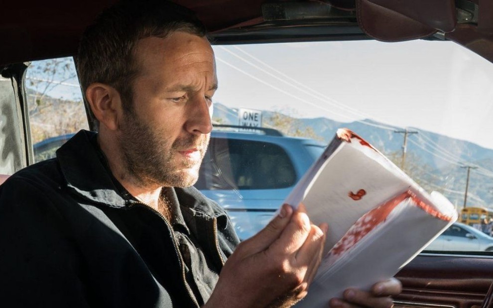 Dentro de um carro, do lado do motorista, o ator Chris O'Dowd folheia um roteiro manchado de sangue na série Get Shorty