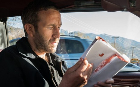 Dentro de um carro, do lado do motorista, o ator Chris O'Dowd folheia um roteiro manchado de sangue na série Get Shorty