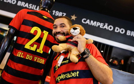 Gil posa ao lado de uma camiseta número 21 com o nome "Gil do Vigor", além de um mascote do clube em mãos
