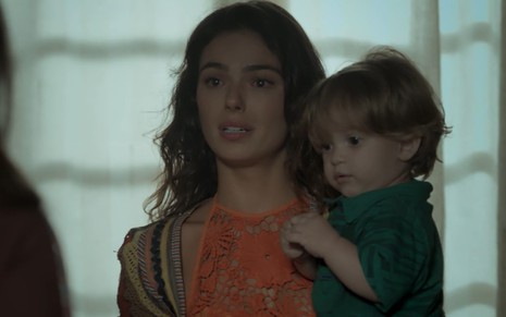 Isis Valverde com cara de choro veste um vestido laranja e carrega um bebê no colo em cena como Ritinha