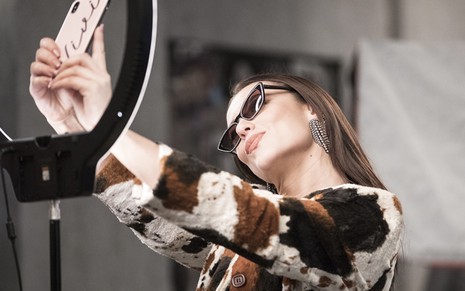 A atriz Paolla Oliveira segura um celular e tira selfie diante de um ring light caracterizada como Vivi em cena de A Dona do Pedaço