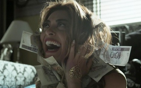 Juliana Paes na cena de A Força do Querer: personagem está caracteriza como Bibi e rola em cima de notas de dinheiro com olhar de impressionada