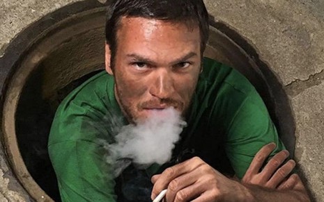 O ator Emilio Dantas caracterizado como Rubinho fuma um cigarro dentro de bueiro nos bastidores de cena de A Força do Querer