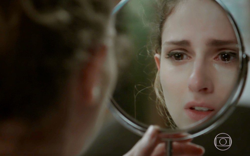 A atriz Carol Duarte olha para seu rosto maquiado em um espelho e estranha durante gravação de cena de A Força do Querer