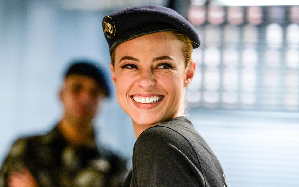 Paolla Oliveira caracterizada como Jeiza em A Força do Querer; com uniforme de policial, personagem sorri para alguém fora do quadro