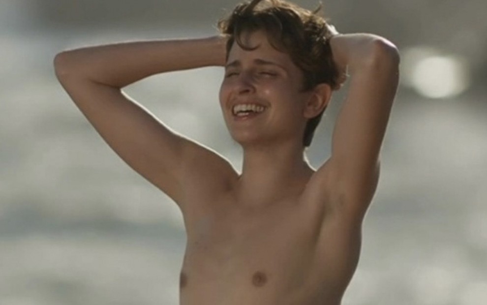 A atriz Carol Duarte em uma praia, leva as mãos à nuca e mostra o peito masculino, em um efeito especial, caracterizada como o Ivan de A Força do Querer
