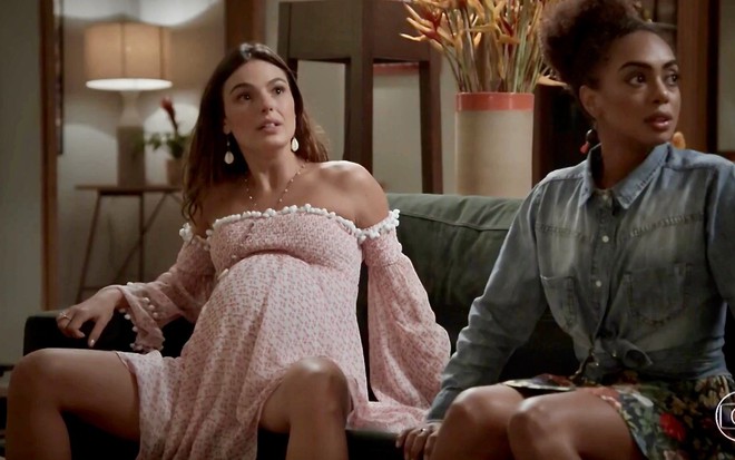 Isis Valverde, com barriga falsa de grávida e sentada com pernas abertas, está sentada ao lado de Dandara Mariana em cena de A Força do Querer