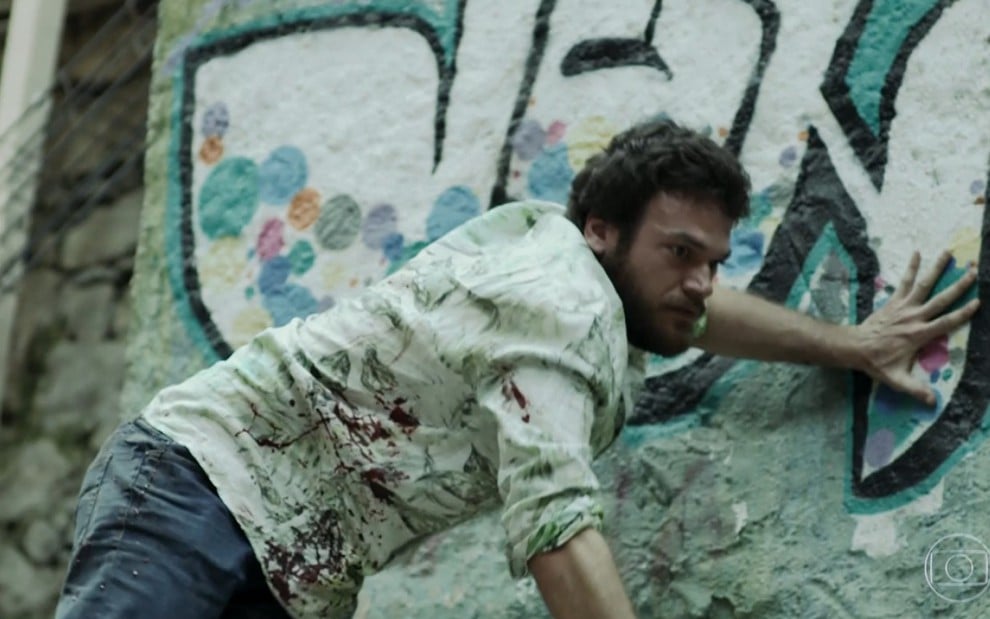 Emílio Dantas grava com camisa manchada de sangue e apoiado em muro em cena de tiro como Rubinho
