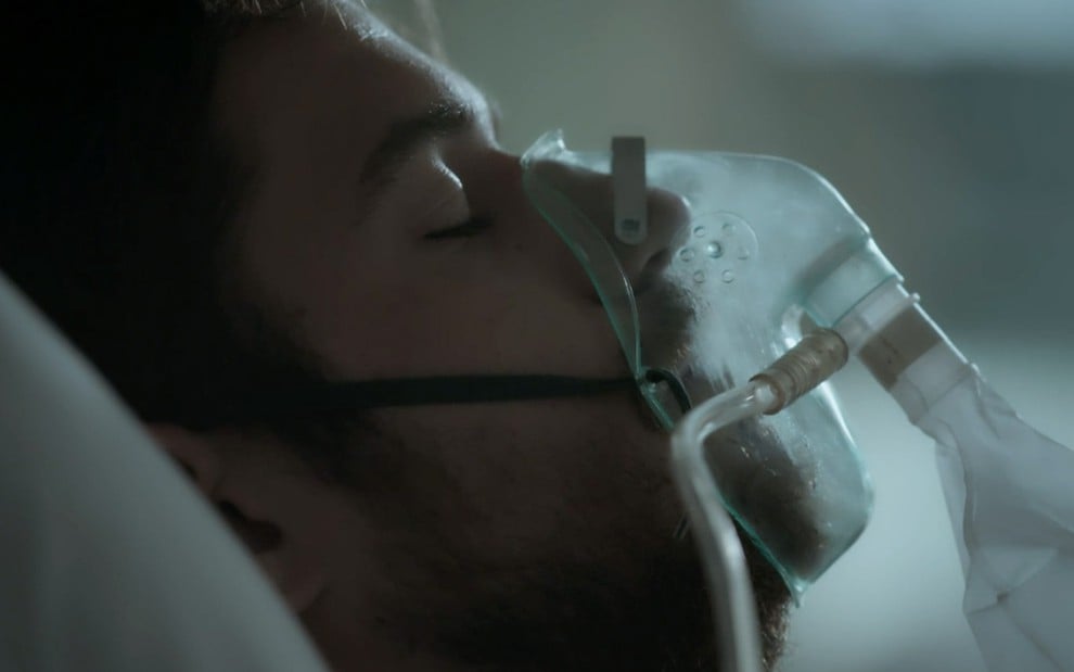 Marco Pigossi caracterizado como Zeca em cena de A Força do Querer: personagem está deitado em cama de hospital e usa equipamento para respirar