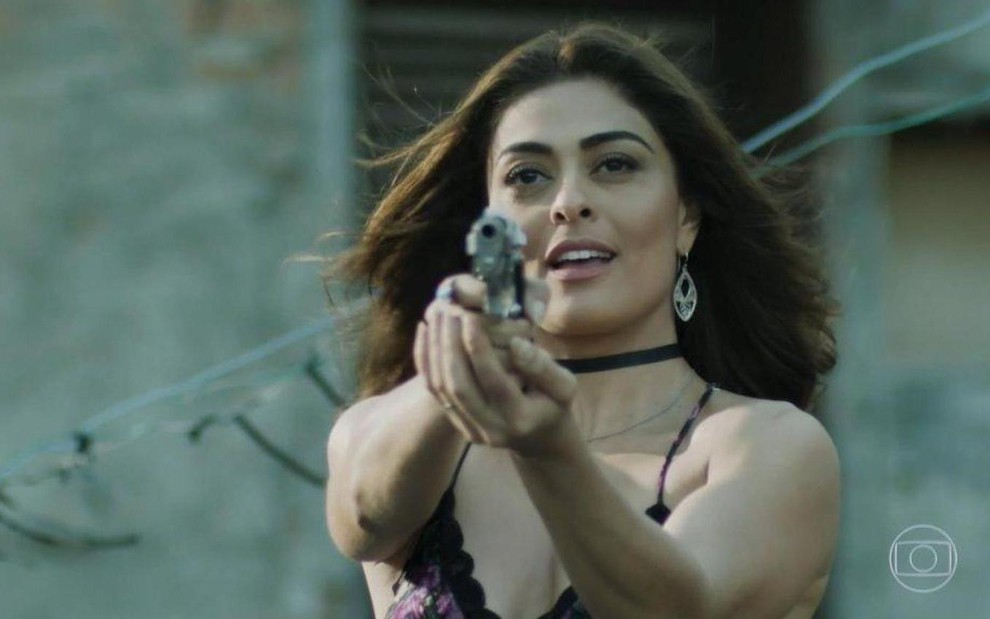 A atriz Juliana Paes segura uma arma e aponta como se fosse atirar em gravação como Bibi da novela A Força do Querer