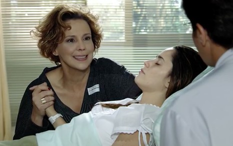 A atriz Ana Beatriz Nogueira como Eva sorri e olha para um homem de costas à direita enquanto segura a mão de Fernanda Vasconcellos, a Ana, que está deitada em um leito de hospital em cena de A Vida da Gente