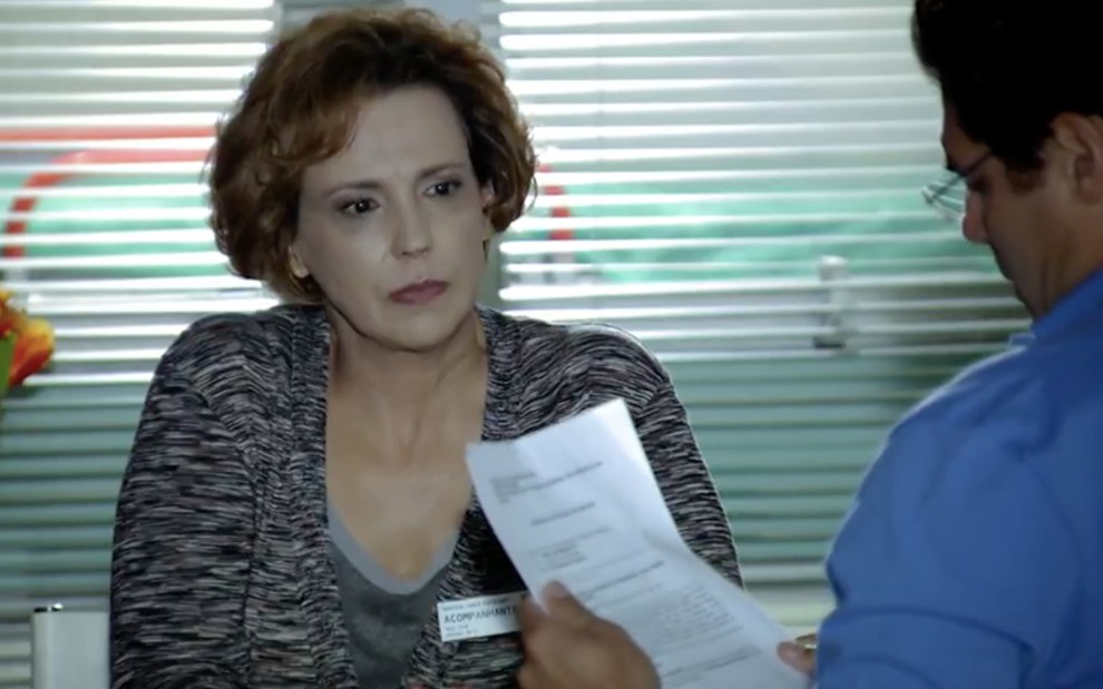 A atriz Ana Beatriz Nogueira está sentada como Eva, na sua frente está Thiago Lacerda, de costas, como Lúcio, segurando um papel com a mão esquerda em cena de A Vida da Gente