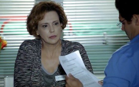 A atriz Ana Beatriz Nogueira está sentada como Eva, na sua frente está Thiago Lacerda, de costas, como Lúcio, segurando um papel com a mão esquerda em cena de A Vida da Gente