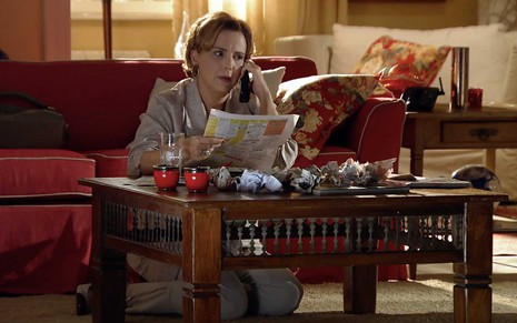 A atriz Ana Beatriz Nogueira está sentado no chão, à frente de uma mesa de centro, com as páginas de classificados na mão esquerda e o telefone na mão direita em cena de A Vida da Gente