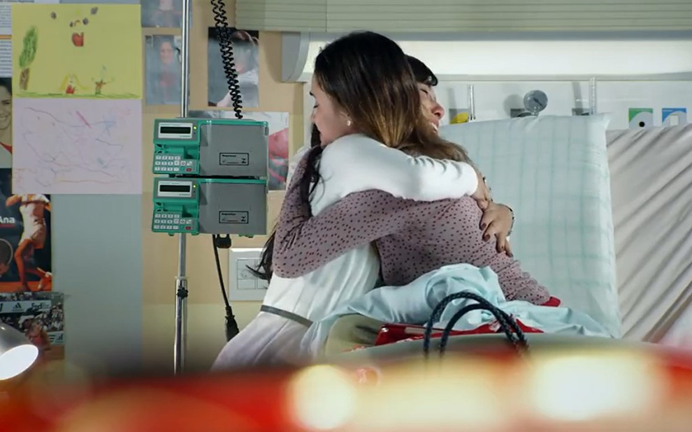 Fernanda Vasconcellos grava sentada na cama de hospital abraçada com Marjorie Estiano, com casaco branco, em A Vida da Gente