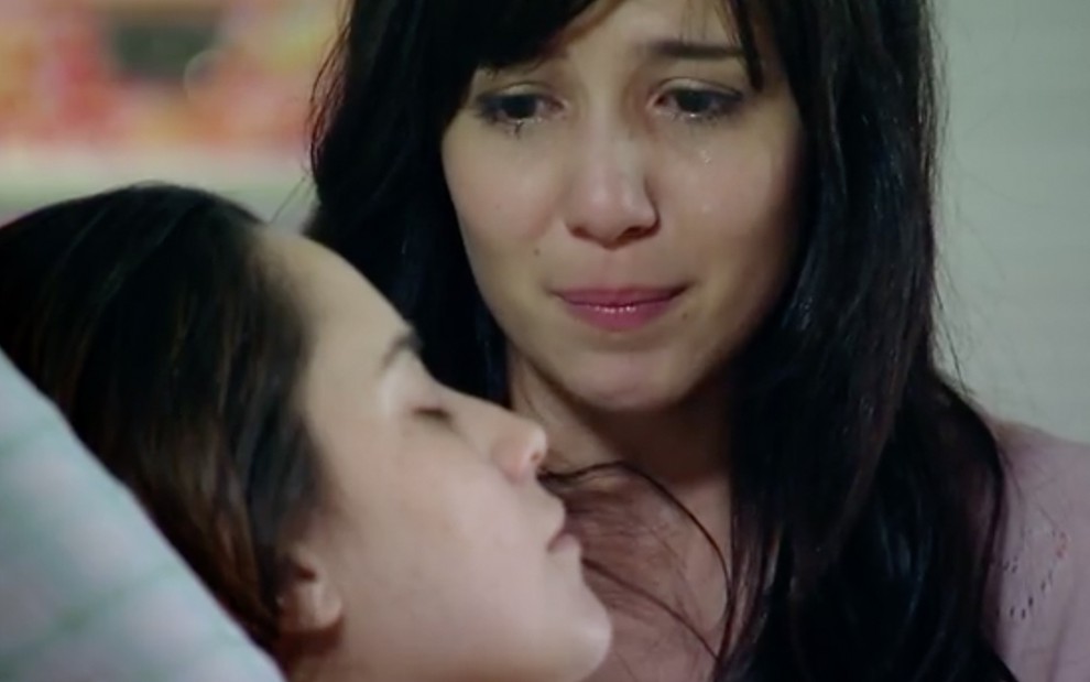 A atriz Fernanda Vasconcellos como Ana deitada em um leito de hospital, de olhos fechados, com a atriz Marjorie Estiano como Manuela sobre o seu corpo com lágrimas nos olhos em cena de A Vida da Gente