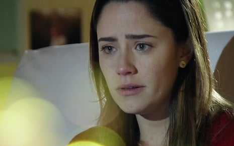 A atriz Fernanda Vasconcellos como Ana está sentada em uma cama de hospital, com lágrimas nos olhos, com um computador em cena de A Vida da Gente