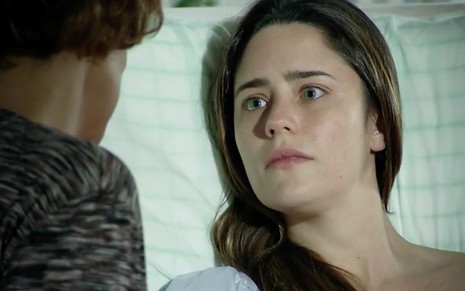 A atriz Fernanda Vasconcellos como Ana tem uma expressão de surpresa enquanto está deitada, de olhos abertos, em uma cama de hospital em cena de A Vida da Gente
