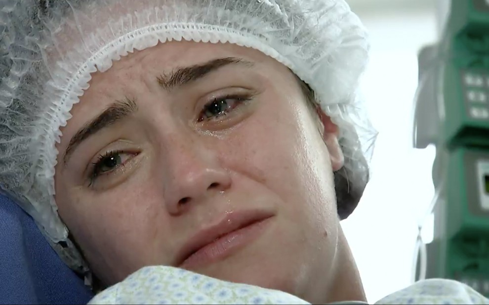 A atriz Fernanda Vasconcellos chora como Ana em uma maca e com uma touca e capote cirúrgicos em cena de A Vida da Gente