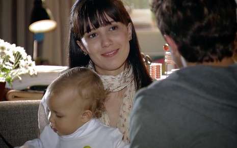 A atriz Marjorie Estiano olha para frente, com um bebê no colo, caracterizada como Manuela em cena de A Vida da Gente