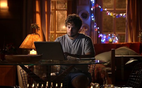 Rafael Cardoso grava sentado escrevendo no computador em cenário iluminado com luzes de Natal como Rodrigo de A Vida da Gente