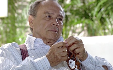 O ator Stênio Garcia como Laudelino está sentado em uma poltrona branca em uma recepção de hospital e segura um terço de madeira escura com as duas mãos, em gesto de oração, em cena de A Vida da Gente
